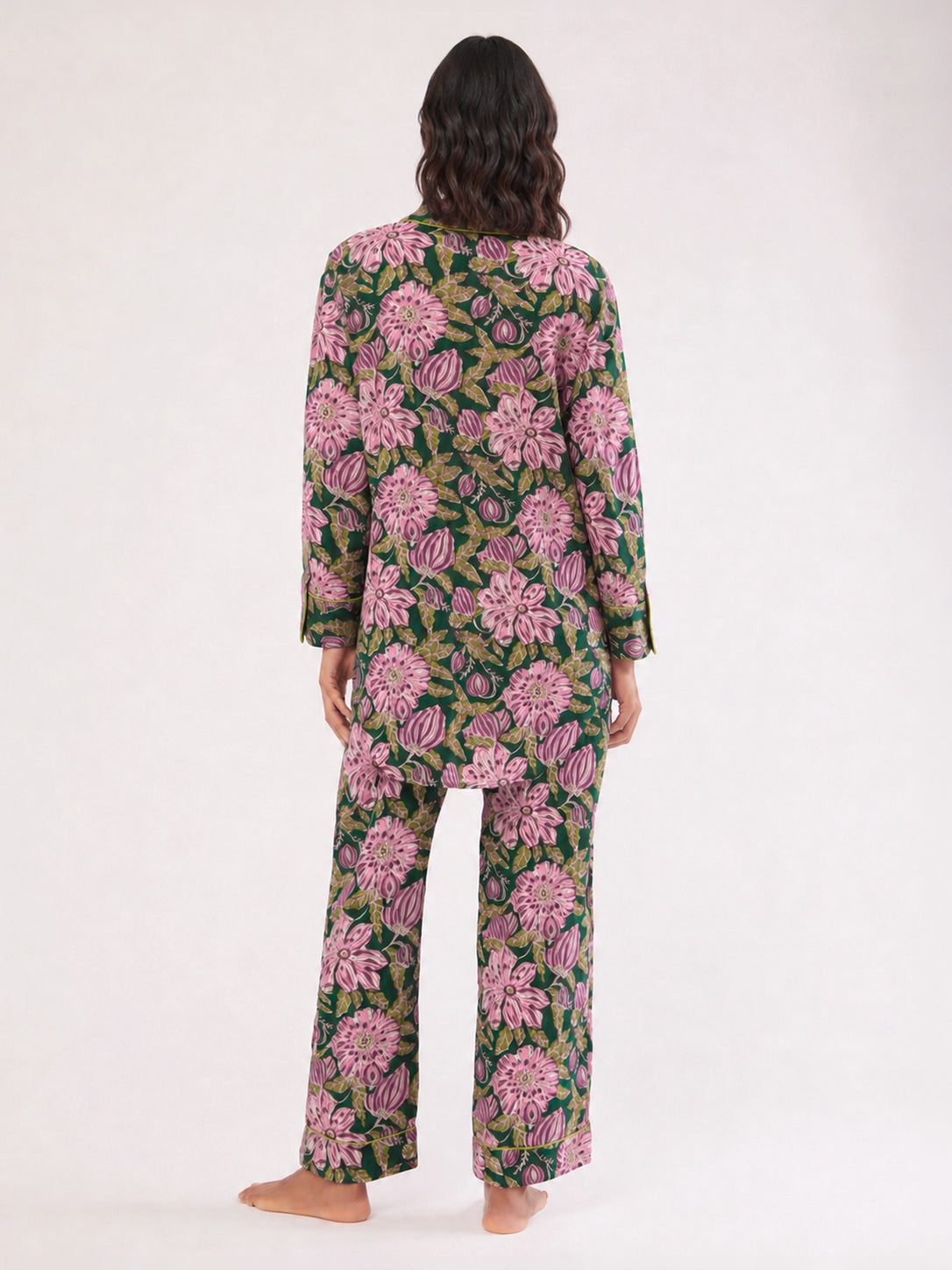 A woman viewed from behind wearing a matching long-sleeve pajama set with a bold pink floral print on a dark green background, standing barefoot against a plain white studio backdrop. ETHICAL FASHION, CRAFTSMANSHIP, SUSTAINABLE FASHION, TRADITIONAL FASHION, ETHNIC FASHION, BOHEMIAN FASHION, MODA ETICA, MODA SOSTENIBILE, TECNICHE ARTIGIANALI, MODA TRADIZIONALE, MODA ETNICA, BOHEMIAN DRESS, MADE IN INDIA, HANDMADE IN INDIA, BLOCK PRINT, FLOWER PAJAMAS, PIGIAMA A FIORI, PIGIAMA DI COTONE, COTTON PAJAMA, SILK P