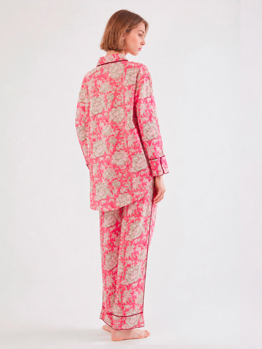 A woman wearing a pink and beige floral pajama set with black piping, shown from the back against a plain white studio background. ETHICAL FASHION, CRAFTSMANSHIP, SUSTAINABLE FASHION, TRADITIONAL FASHION, ETHNIC FASHION, BOHEMIAN FASHION, MODA ETICA, MODA SOSTENIBILE, TECNICHE ARTIGIANALI, MODA TRADIZIONALE, MODA ETNICA, BOHEMIAN DRESS, MADE IN INDIA, HANDMADE IN INDIA, BLOCK PRINT, FLOWER PAJAMAS, PIGIAMA A FIORI, PIGIAMA DI COTONE, COTTON PAJAMA, SILK PAJAMA, PIGIAMA DI SETA, VESTAGLIA, NIGHTWEAR, SLEEPWE