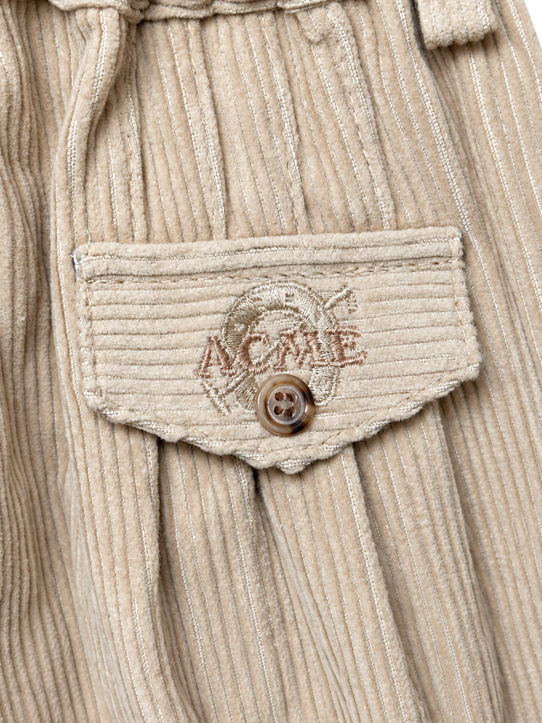 Close-up of a beige ribbed corduroy garment featuring a front pocket with a scalloped edge, a brown button, and embroidered ACME branding. Argentine Bombacha Pants, horseback riding, traditional gaucho, Argentine cowboy attire. Soft and breathable fabrics, perfect pants for all-day wear. With a loose fit and elasticated waistband, they allow for plenty of movement, while the high waist provides extra coverage. Everyday look, perfect for both urban and countryside adventures. Cotton Corduroy, KIDS, PANTS, KI