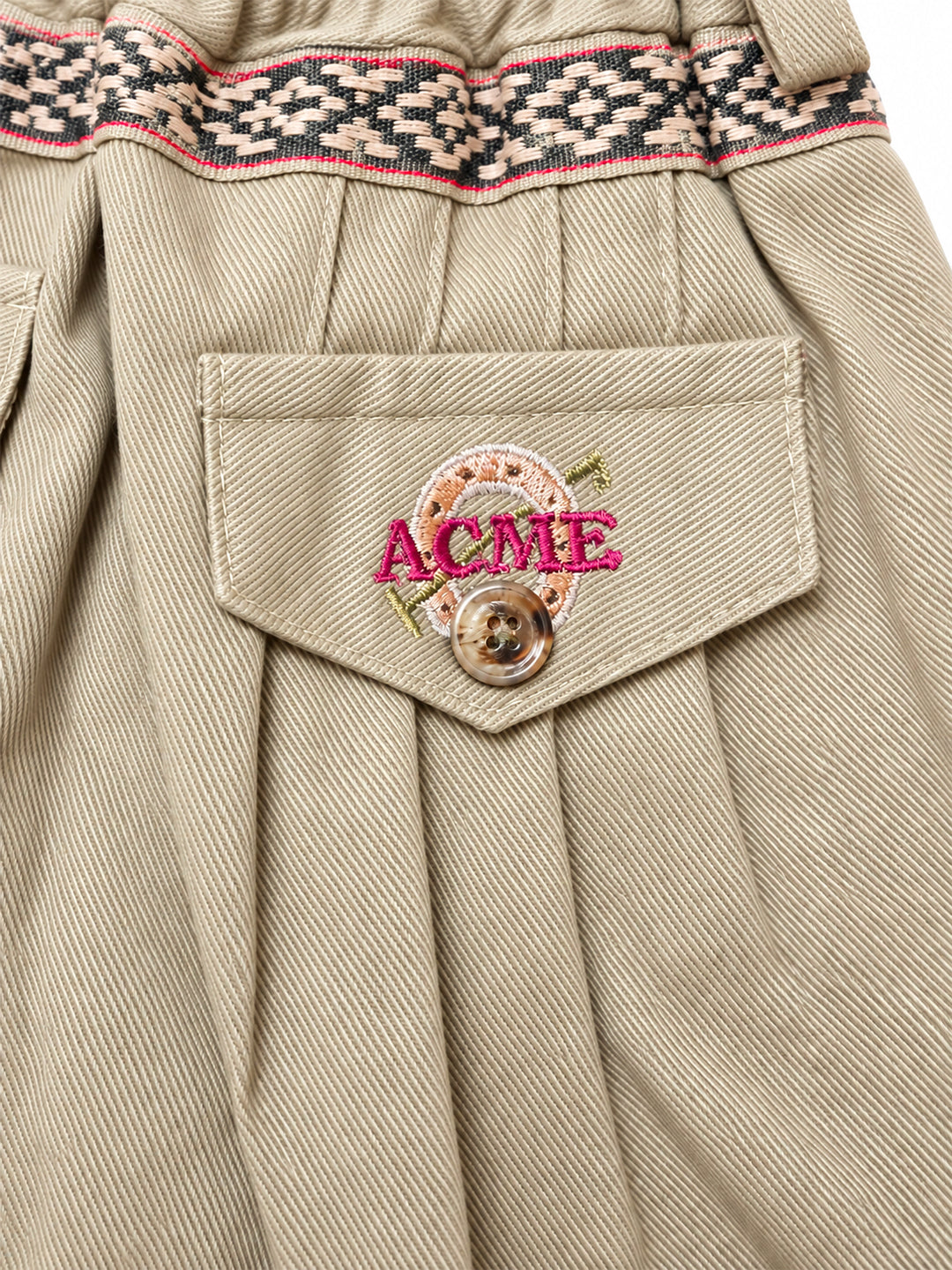 Close-up of pleated beige trousers with a decorative patterned waistband and a front pocket featuring a pink embroidered 'ACME' patch with a button closure. Argentine Bombacha Pants, horseback riding, traditional gaucho, Argentine cowboy attire. Soft and breathable fabrics, perfect pants for all-day wear. With a loose fit and elasticated waistband, they allow for plenty of movement, while the high waist provides extra coverage. Everyday look, perfect for both urban and countryside adventures. Cotton Corduro