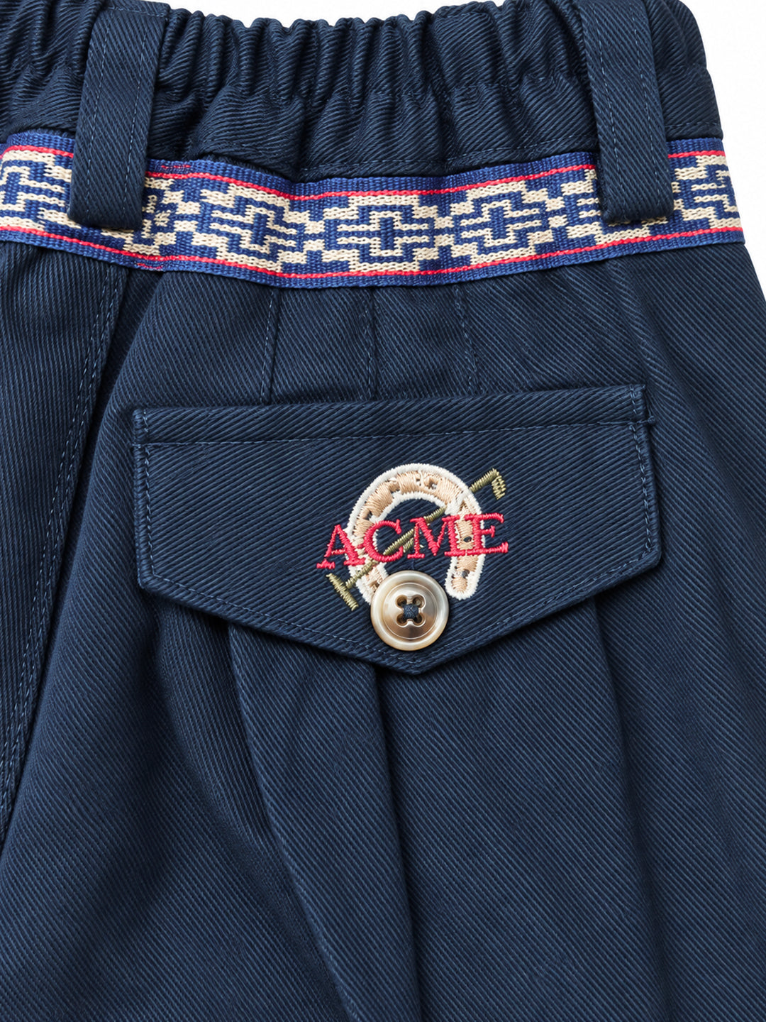 Close-up of navy blue pleated trousers featuring a decorative patterned waistband and a front pocket with a button and embroidered red 'ACME' Argentine Bombacha Pants, horseback riding, traditional gaucho, Argentine cowboy attire. Soft and breathable fabrics, perfect pants for all-day wear. With a loose fit and elasticated waistband, they allow for plenty of movement, while the high waist provides extra coverage. Everyday look, perfect for both urban and countryside adventures. Cotton Corduroy, KIDS, Ptext.