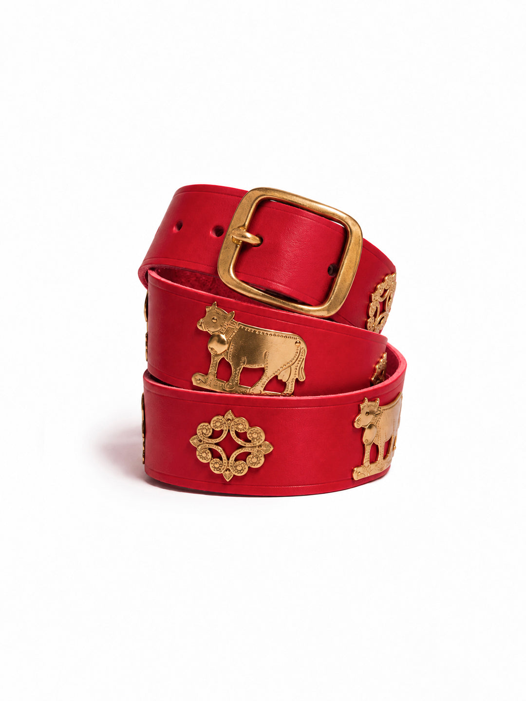 A red leather belt with gold-tone metal accents, including a square buckle, decorative medallions, and cow motifs, coiled on a white background. The Alpine Belt, Appenzeller belt, made in Switzerland, handmade, mountain belt, leather belt, cintura di pelle, cintura con le mucche, stelle alpine, cintura svizzera, cintura tirolese, cintura ampezzana, traditional belt, cow belt, kids belt, artisanal kids belt, artisanal belt, cinture da bambini, cinture artigianali.