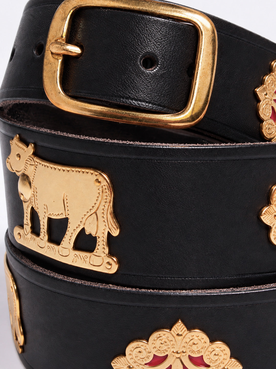 A close-up of a black leather belt with ornate gold-tone metal hardware, including a large rectangular buckle, a cow-shaped emblem, and decorative floral motifs with small red accents. The Alpine Belt, Appenzeller belt, made in Switzerland, handmade, mountain belt, leather belt, cintura di pelle, cintura con le mucche, stelle alpine, cintura svizzera, cintura tirolese, cintura ampezzana, traditional belt, cow belt, kids belt, artisanal kids belt, artisanal belt, cinture da bambini, cinture artigianali.